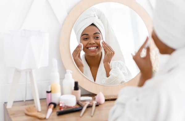 Pretty african american woman removing makeup, cleaning face with cotton pads, looking in mirror while sitting at dressing table