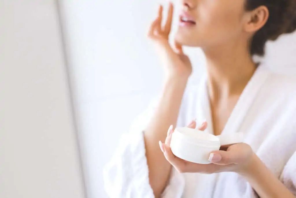 Woman in white bathrobe applying face cream from a jar