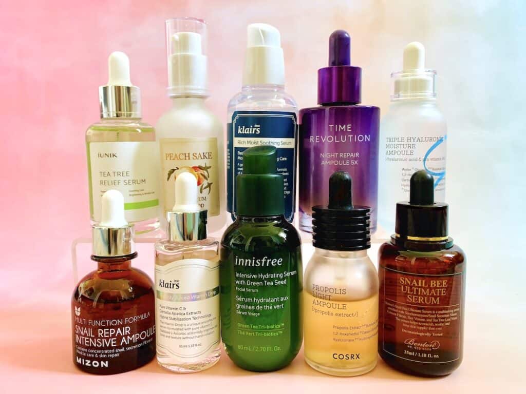 Best Korean serums from Mizon, Klairs, innisfree, COSRX, Benton, iUNIK, Skinfood and Missha: 5 serums in front and 5 serums on risers in back. On pink background.